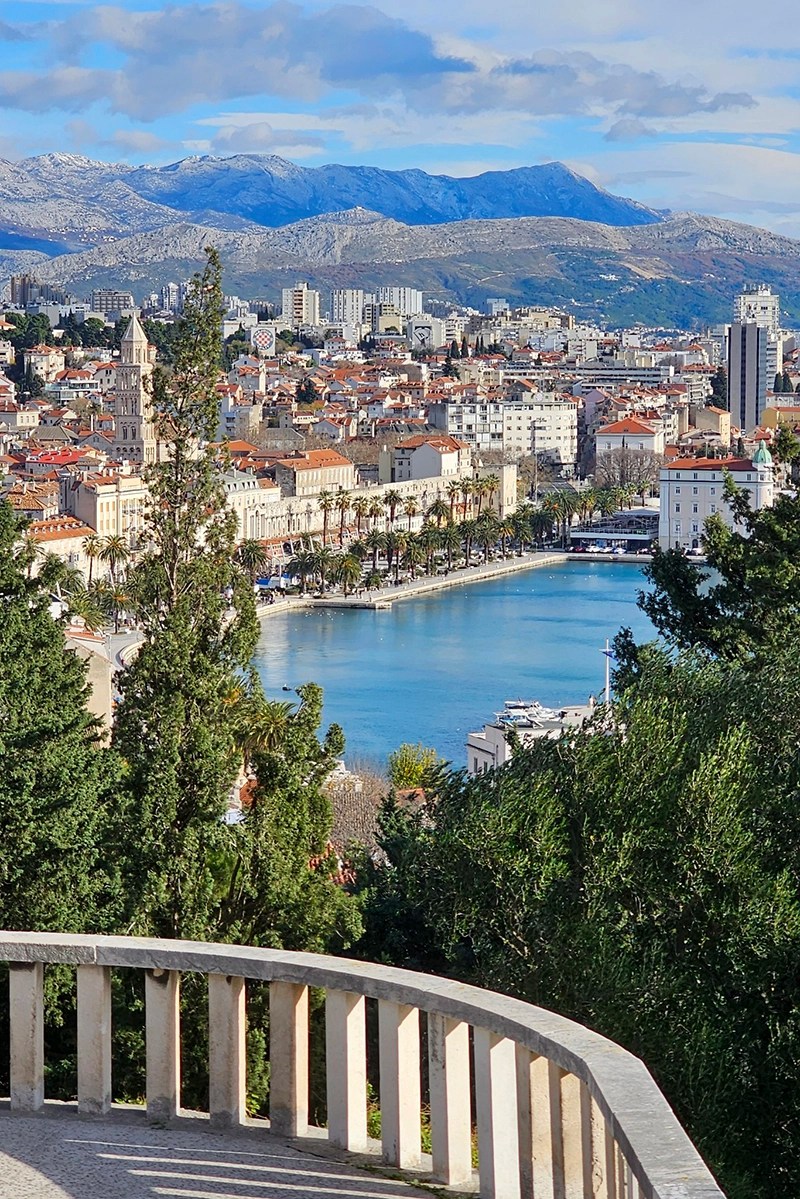 Vista panorâmica da cidade costeira de Split, Croácia, com edifícios de telhados vermelhos, um calçadão ladeado por palmeiras e montanhas ao fundo sob um céu parcialmente nublado.