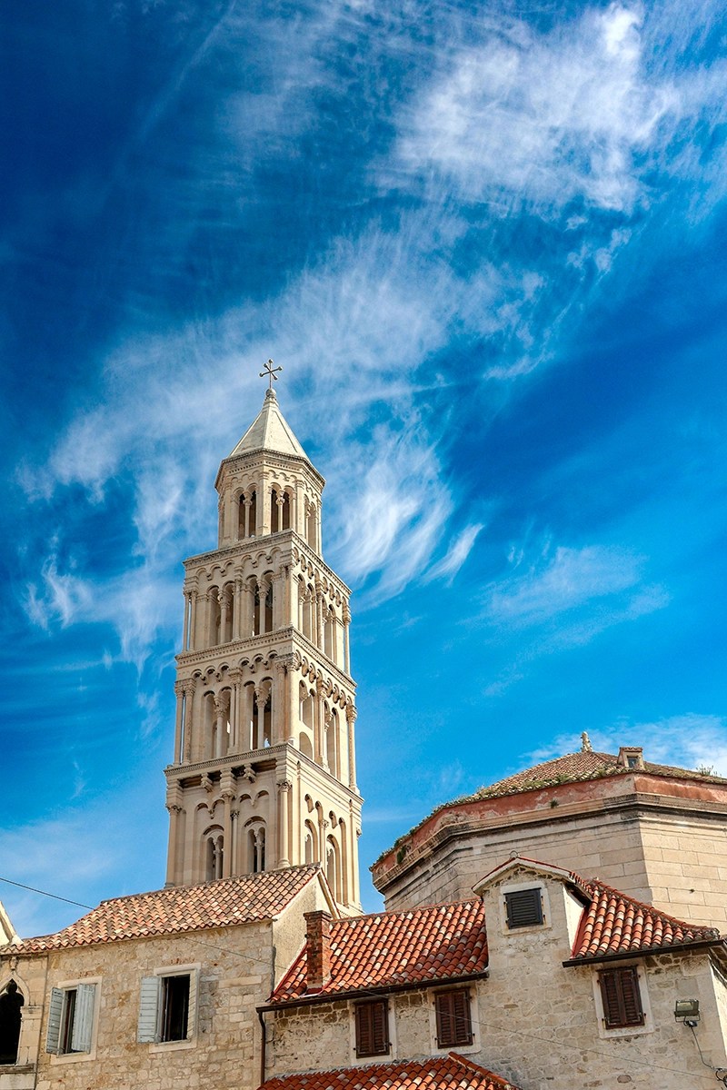 A torre sineira da Catedral de São Dômnio em Split, Croácia, com arcos ornamentados, ergue-se acima dos telhados de telha vermelha sob um céu azul vibrante com nuvens finas.