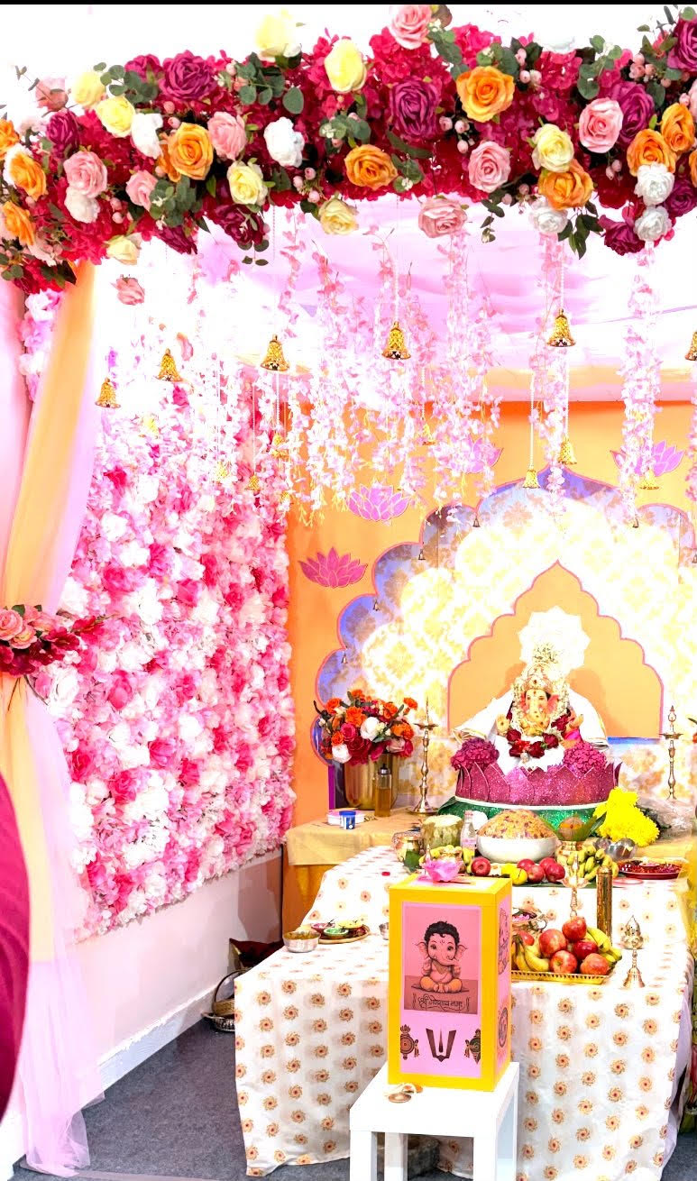 The pandal, built inside the host family's garage: a canopy of roses, cascading blossoms and golden bells, lotus motifs on the walls, and Lord Ganesha seated beneath the arch with fruit and offerings at his feet