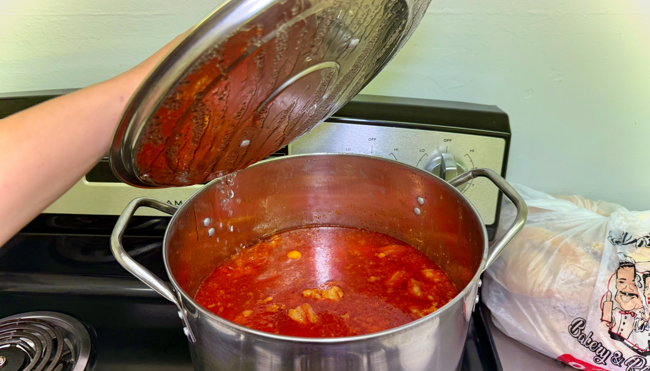 Hand holding open the lid to a large pot of red soup that has condensation. For context, to the right of the soup pot is a bag of bread with an animated mexican chef couple as the logo. 