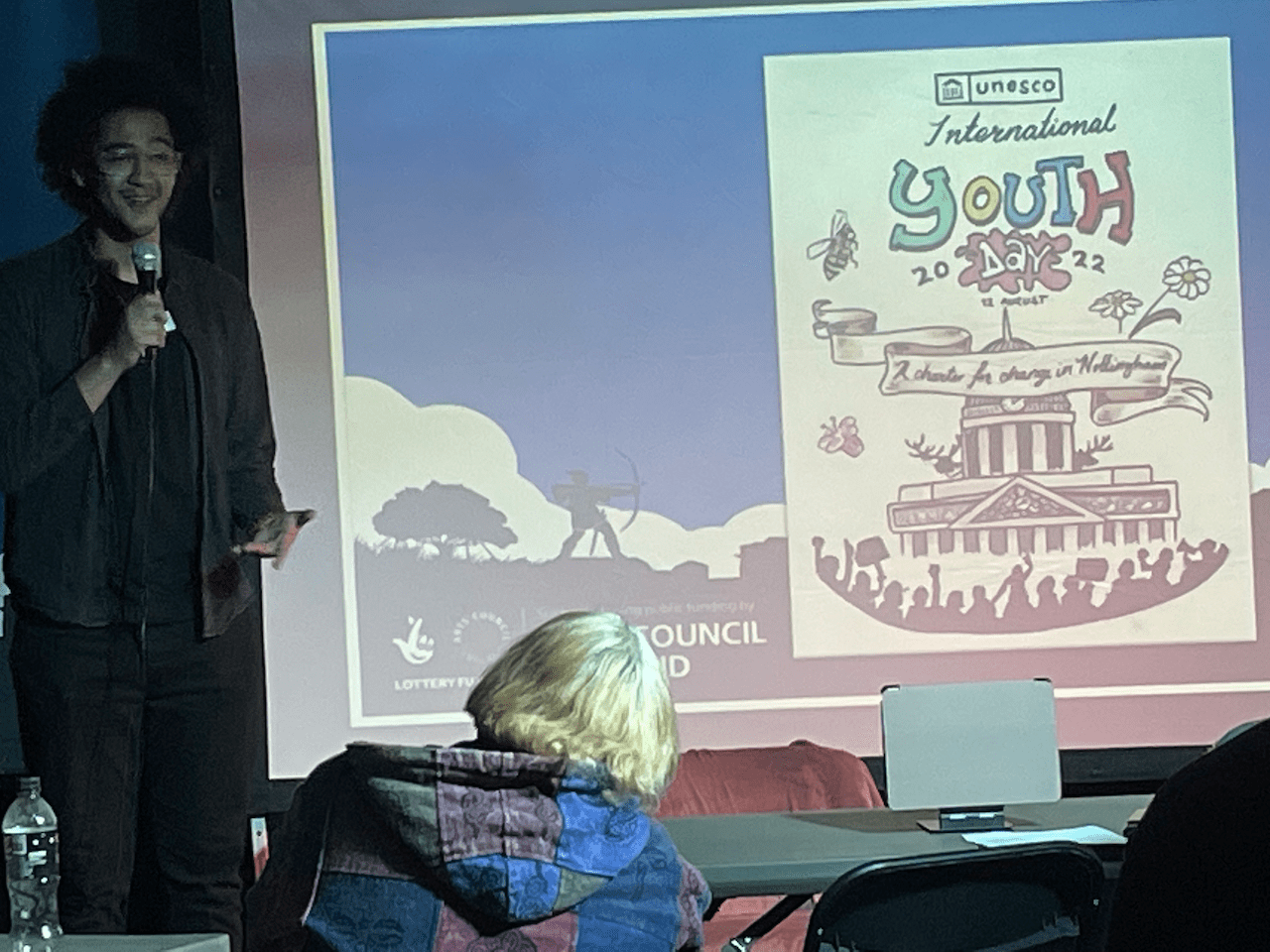A performance poet standing next to a screen with an illustration saying 2022 international youth day