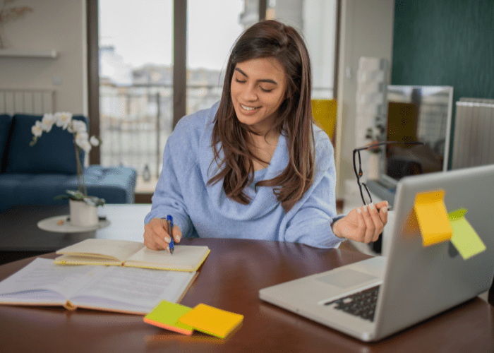 A light skinned woman with long brown hair is wearing a blue cowl neck sweater writing in a journal. Her glasses are in her left hand and her laptop is in the foreground. She is smiling at her book.
