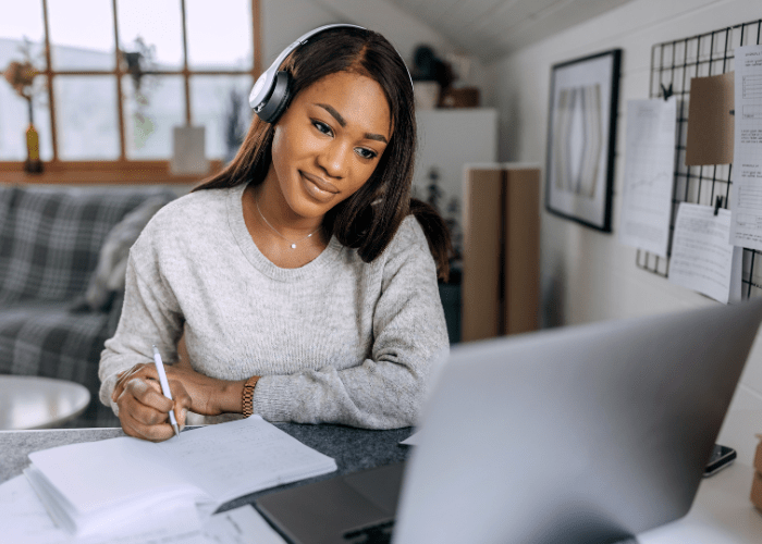 A dark skinned woman with long brown hair is wearing a grey sweater and headphones looking at a computer screen while she takes notes.