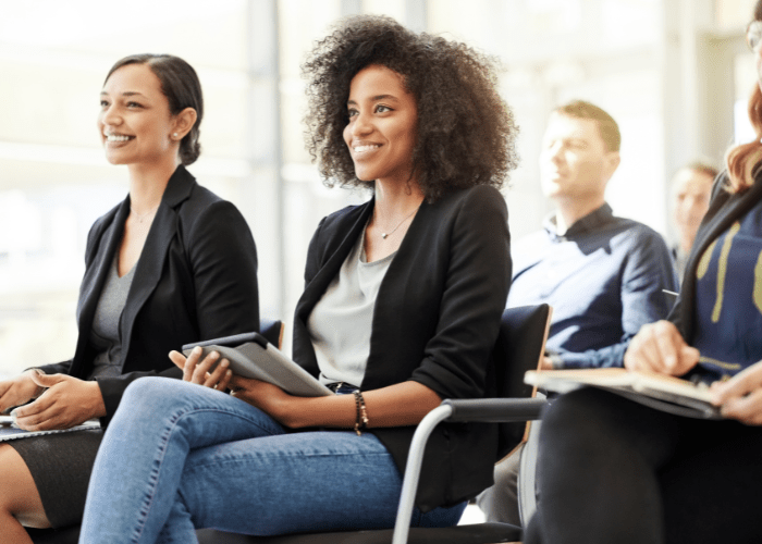 A group of people are seated in a well-lit room wearing business casual clothing. A dark-skinned woman with curly brown hair is seated at the forefront of the room and a light-skinned woman with a bun in her hair is seated next to her. A light-skinned man is seated behind them.