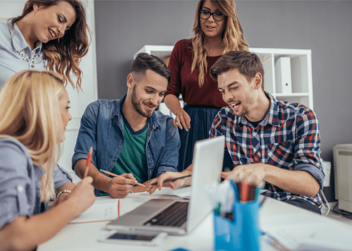 A group of casually dressed, light-skinned men and women gather around a work table with a laptop in the forefront. they are smiling and look to be in agreement about the work in front of them.