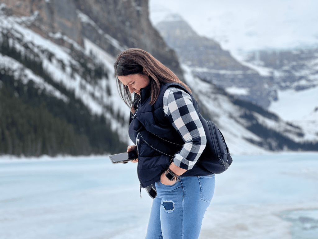 Heather Deveaux is wearing a black vest and black and white check shirt with jeans and is looking toward the ground with ice and snow-covered mountains in the background.