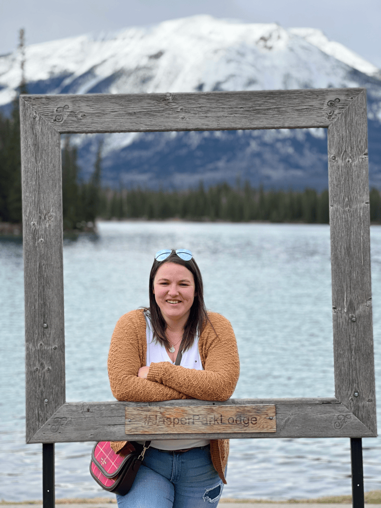 Heather Deveaux leans on a wooden frame that reads Jasper with a lake and a snow-covered mountain appearing in the background. She wears a yellow cardigan and white tshirt. Her arms are crossed in front of her as she leans on the frame.