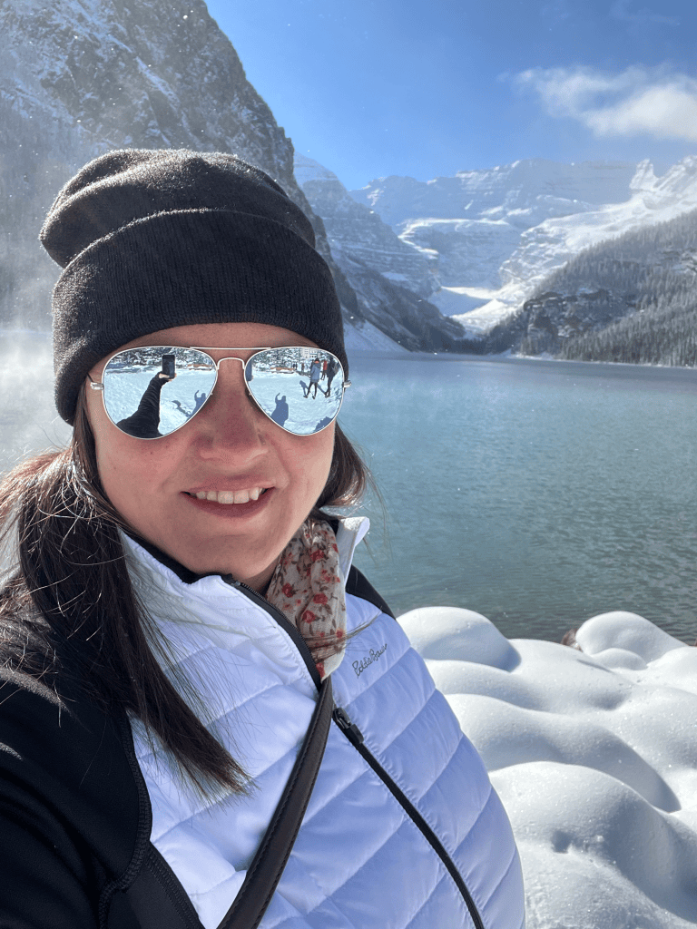 Heather Deveaux wears sunglasses and a winter coat and hat and is pictured in front of Lake Louise with snow-covered rocks and mountains in the background.