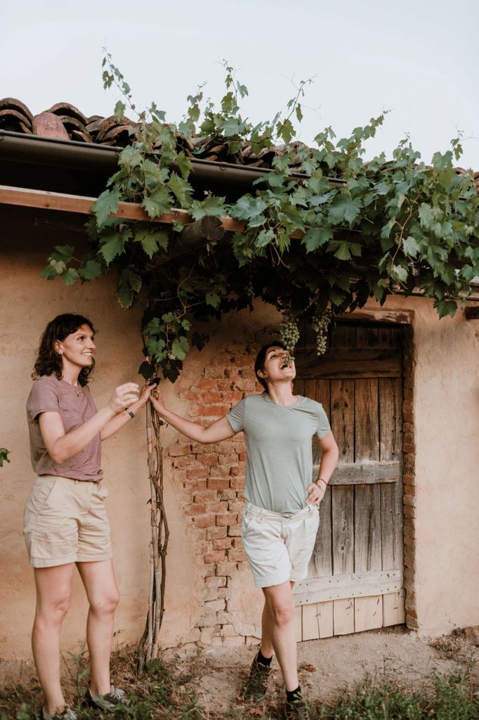 Two women standing near a grapevine with a rustic building in the background, enjoying their time together in a vineyard.