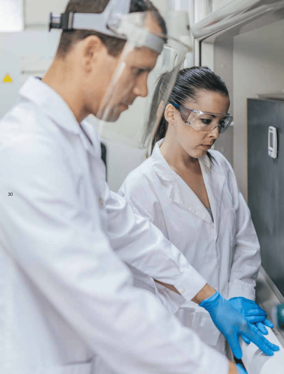Two scientists wearing lab coats and safety goggles, one applying gloves, working in a laboratory setting.