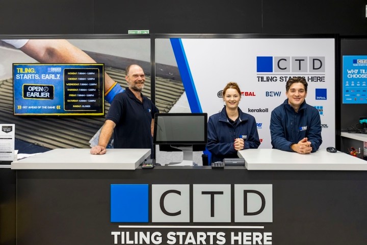 Three employees standing behind a customer service desk at a tiling store, with promotional signage and a digital display behind them.