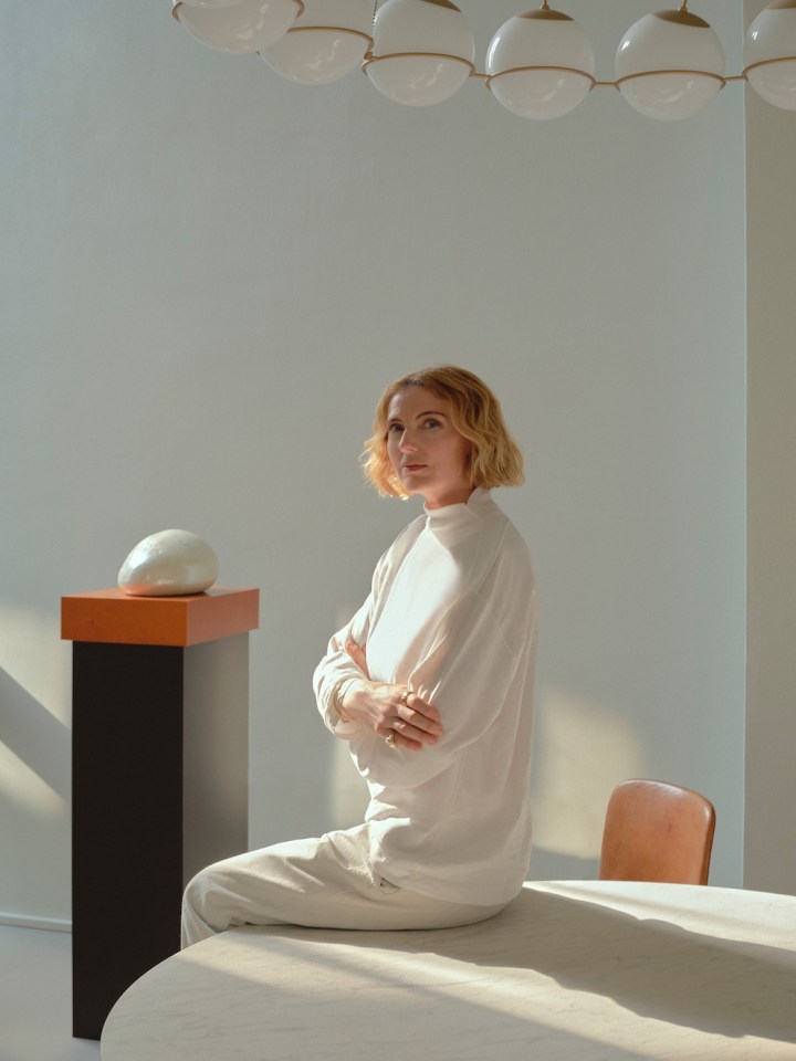 A woman with short, wavy hair sits at a round table, wearing a white outfit. She has her arms crossed and looks thoughtfully at the camera, with soft lighting illuminating her face. In the background, there is a modern lamp with spherical shades and an abstract sculpture on a pedestal.