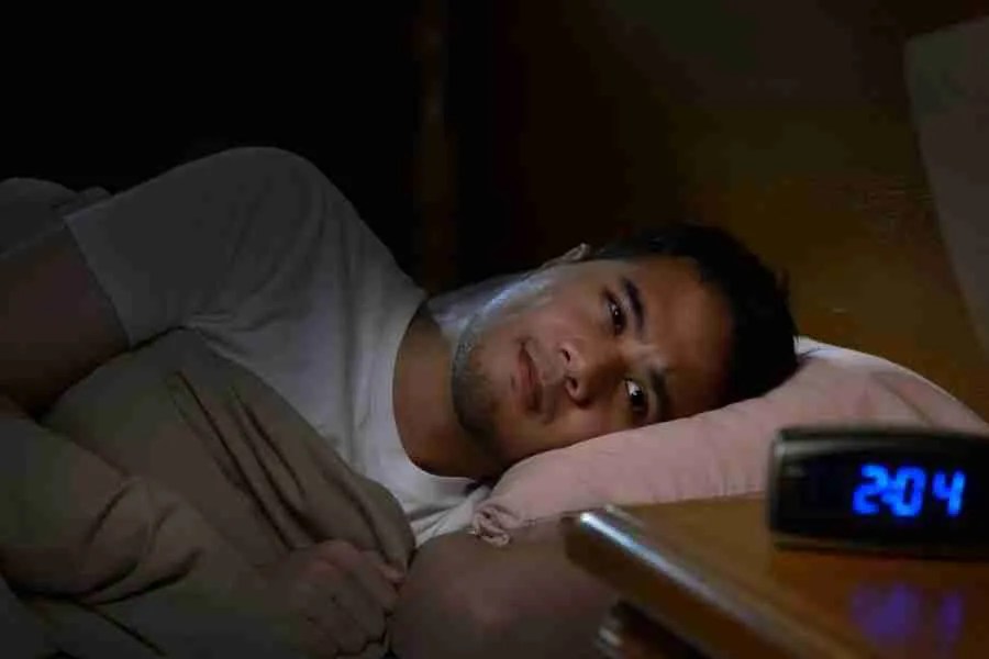 A person lies in bed, awake at night, pondering anxiously under a beige blanket on a pink pillow. As the digital clock reads 2:04, they wonder, Do I have anxiety? When is it time to seek help for anxiety therapy?.