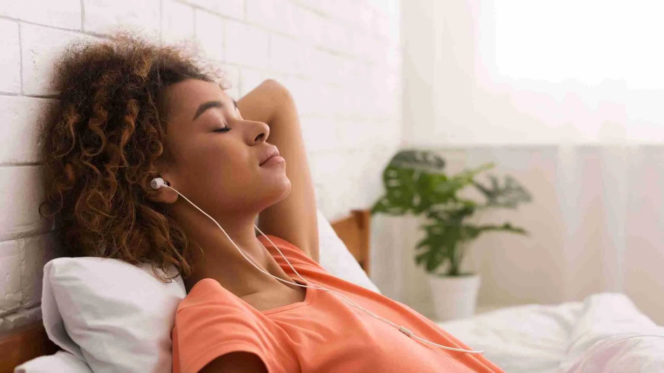 A person with curly hair rests against a pillow, eyes closed, embraced by the soothing effects of music therapy through earphones. Dressed in an orange shirt, they find tranquility as a green plant adds calm to the softly lit room.