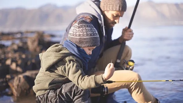 A man and a child sit by the water, both dressed warmly in the spirit of conscious parenting. The child, wearing a beanie and scarf, holds a fishing rod and intently focuses on an object in his hand. The man also holds a fishing rod. Behind them, mountains and shimmering water unfold.