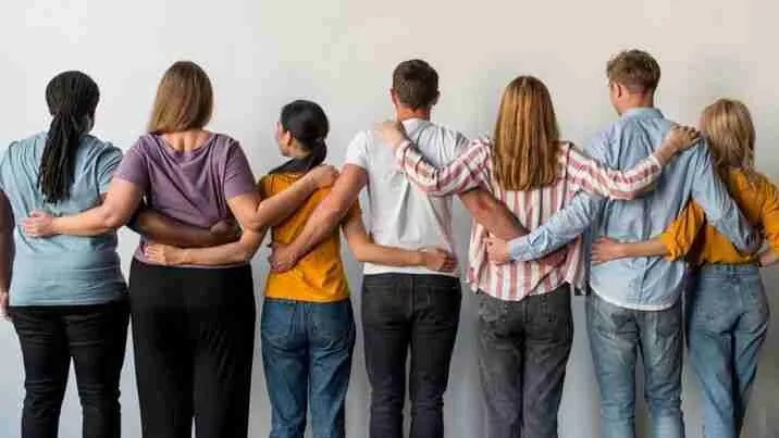 A group of seven diverse people embraces the spirit of finding community, standing in a row with arms around each others shoulders. They face away from the camera, wearing casual clothing against a plain, light-colored background, symbolizing unity and friendship.