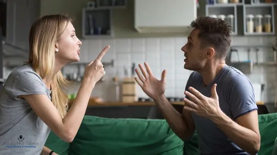 A woman and a man are sitting on a green couch in the kitchen, locked in a heated argument. The woman points her finger while the man gestures animatedly. Both appear frustrated, perhaps highlighting the strain that ADHD and divorce can place on relationships, with kitchen shelves looming in the background.