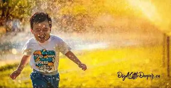 A young boy joyfully runs through a sunlit spray of water on a grassy field, offering a quick stress relief to anyone watching. He wears a colorful t-shirt and smiles broadly, with the golden light creating a warm, cheerful atmosphere.