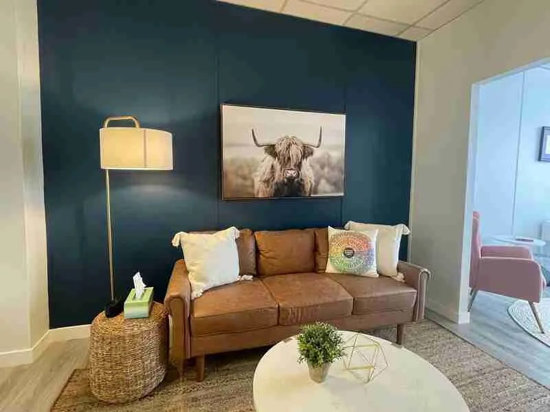 A cozy living room with a brown leather sofa adorned with cushions, perfect for virtual therapy in Ontario. A large floor lamp stands beside it on a woven side table. The dark blue wall features a framed photo of a highland cow, while the round coffee table holds decor items.