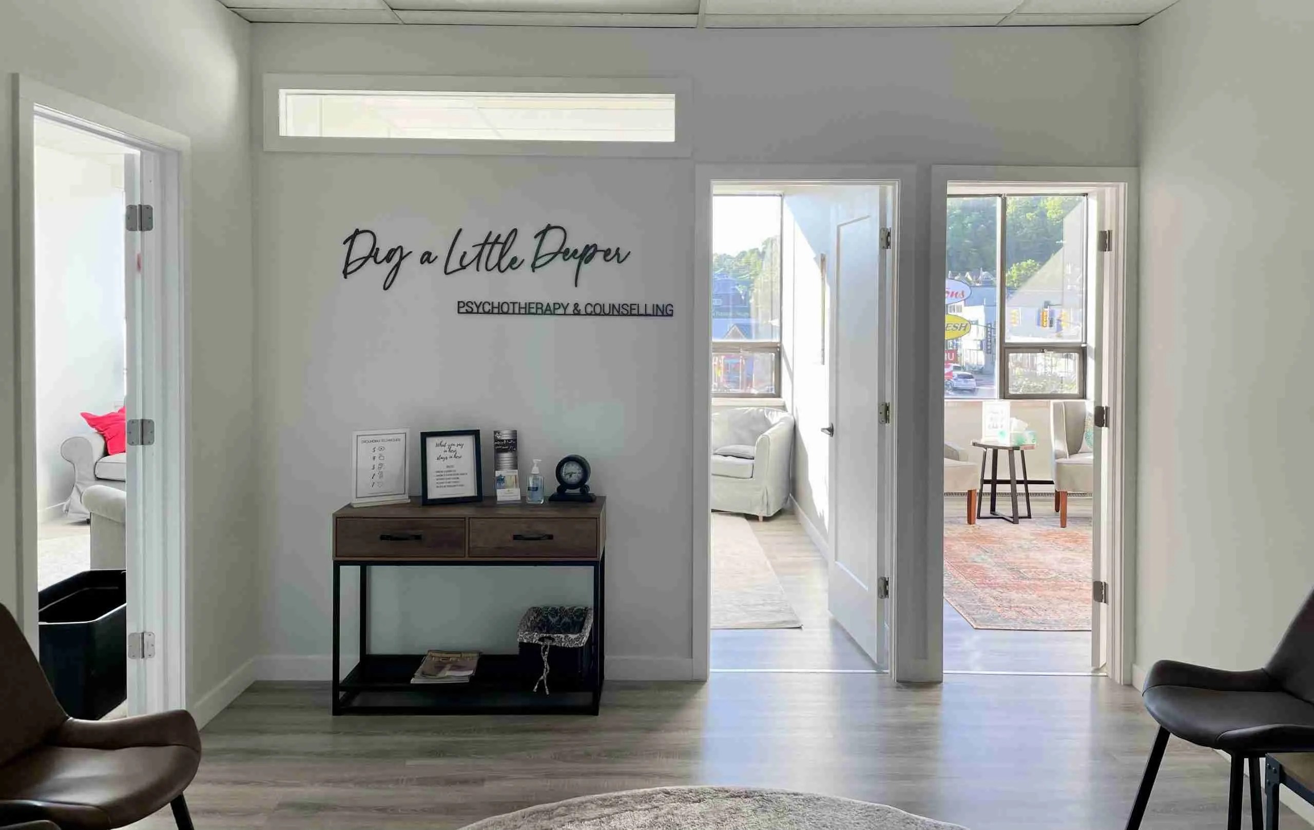 A bright office interior with two open doors revealing lounge areas. A wooden console table against the wall displays framed items and a clock. Text on the wall reads Dig a Little Deeper, In-Person Therapy Ontario. Natural light fills the space.