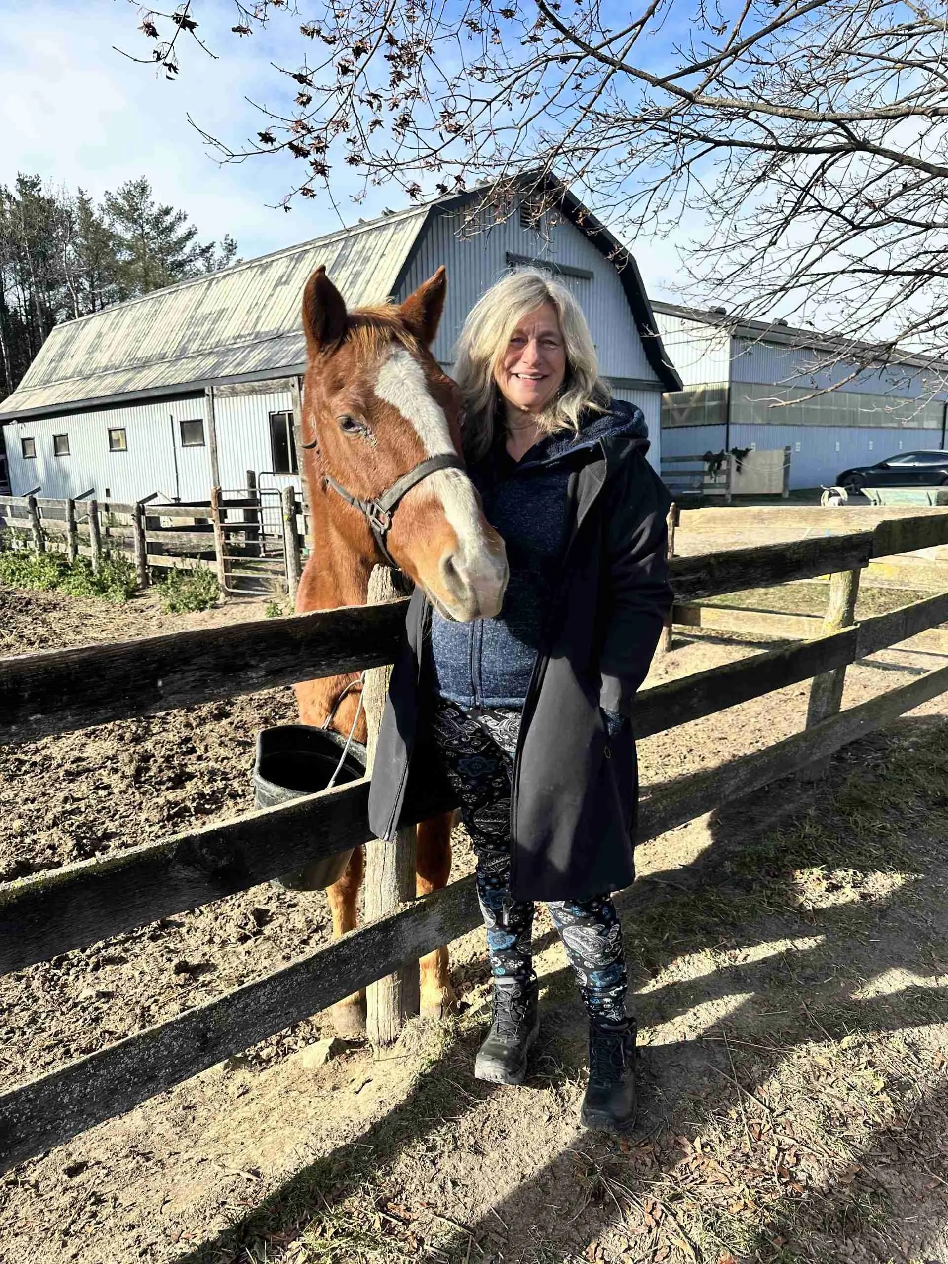 A woman stands outdoors next to a brown horse near a wooden fence, embodying the serenity often sought in Caledon Therapy. She wears a black coat and patterned leggings, with a barn and trees framing the peaceful Ontario backdrop on this clear day.