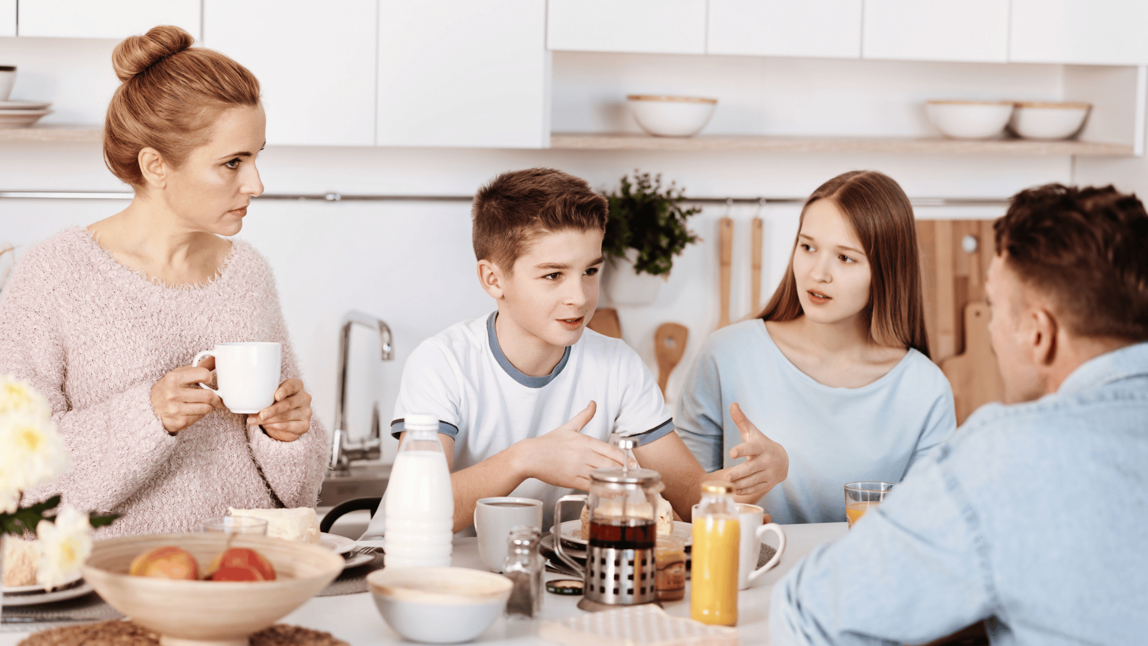 A family gathered at the kitchen table navigates their morning routine with understanding and compassionate boundaries. The woman cradles a mug, while the teenage boy gestures animatedly, a girl listens intently, and a man engages warmly. Juice and pastries adorn the scene.