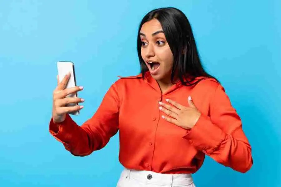 A woman in a red shirt stands against a blue background, looking surprised as she holds a smartphone in her hand. Her mouth is open in astonishment, her other hand placed on her chest, perhaps just having read about cultural stigma and ADHD within South Asian communities.