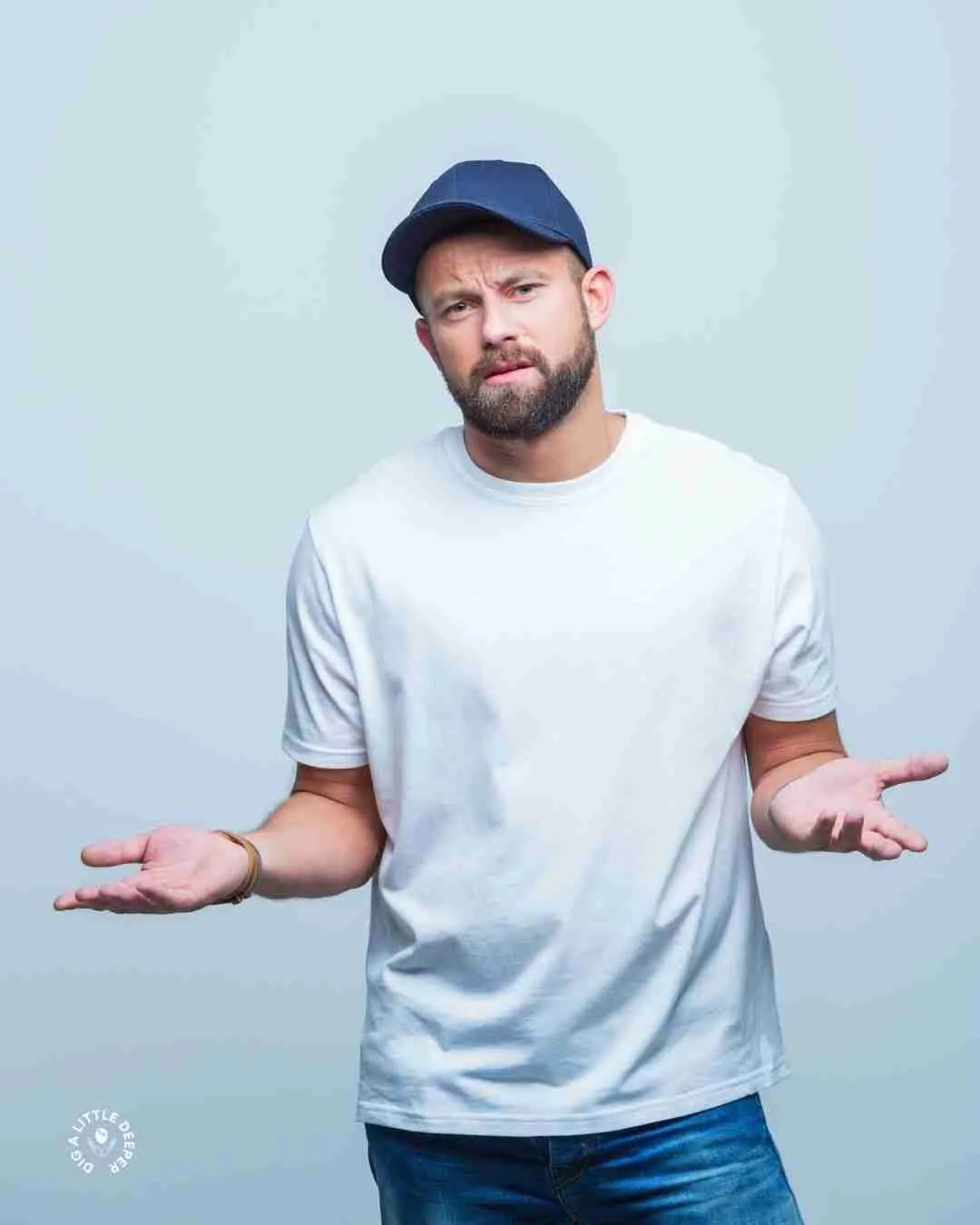 A man with a beard wearing a navy blue cap, white t-shirt, and jeans stands against a plain light background, shrugging with both hands raised and an uncertain expression—like someone navigating ADHD treatment Ontario options.