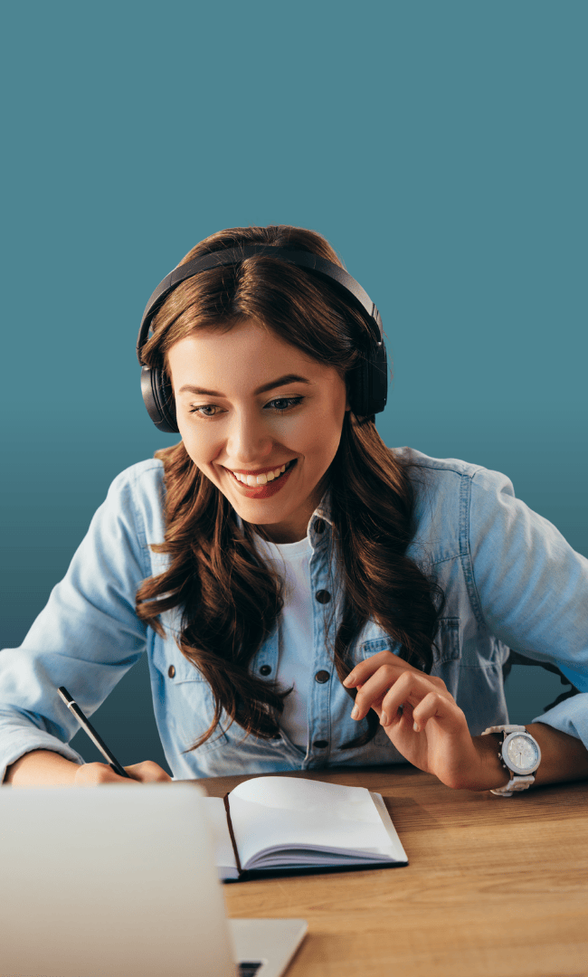 A young woman with long brown hair wearing headphones and a denim shirt is smiling while taking notes in a notebook, sitting at a desk with a laptop, against a plain teal background.