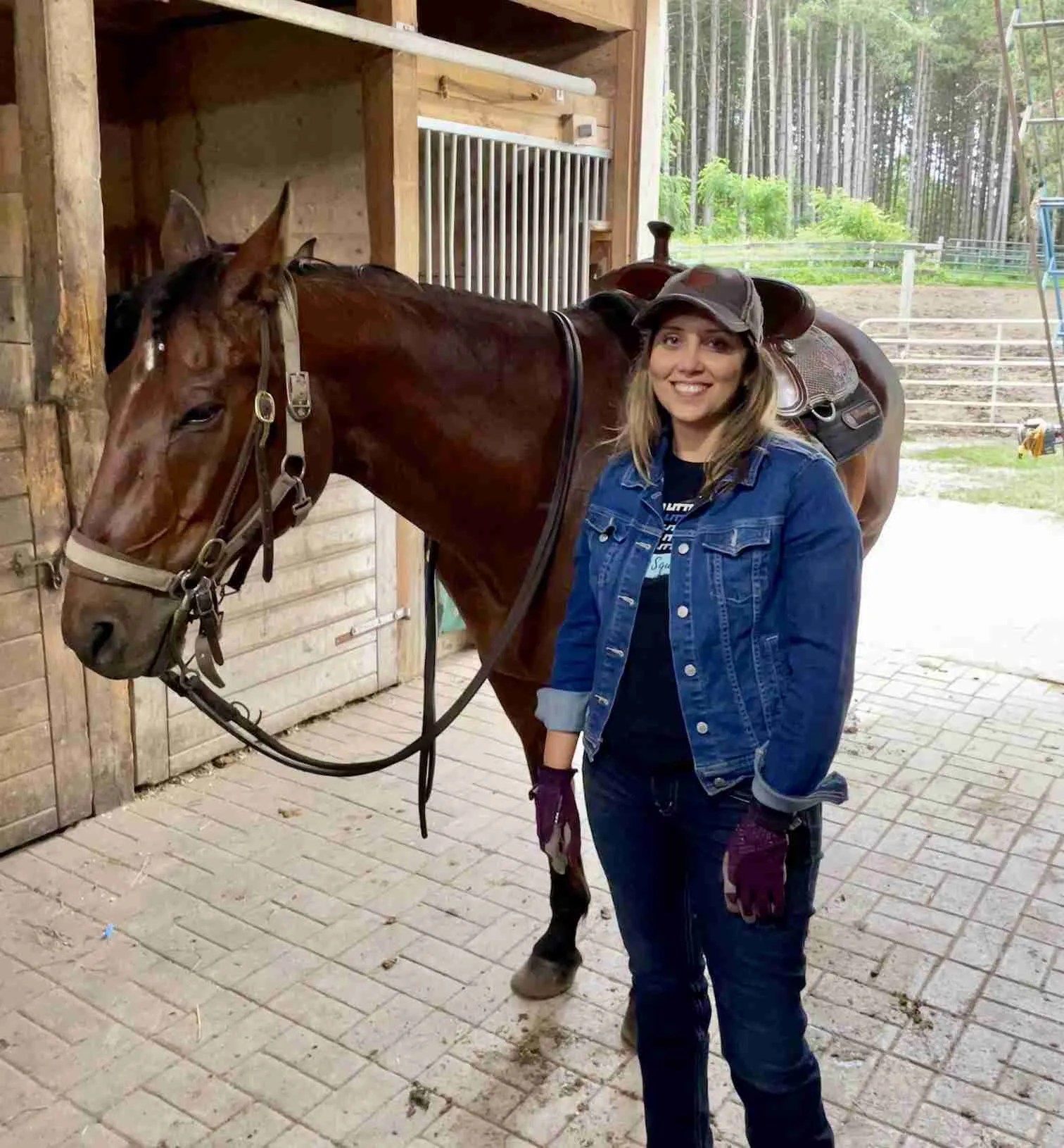 A woman wearing a denim jacket, gloves, and a cap stands next to a saddled brown horse inside a stable. She is smiling, reflecting the positive impact of equine therapy for anxiety. Trees are visible outside through an open stable door.