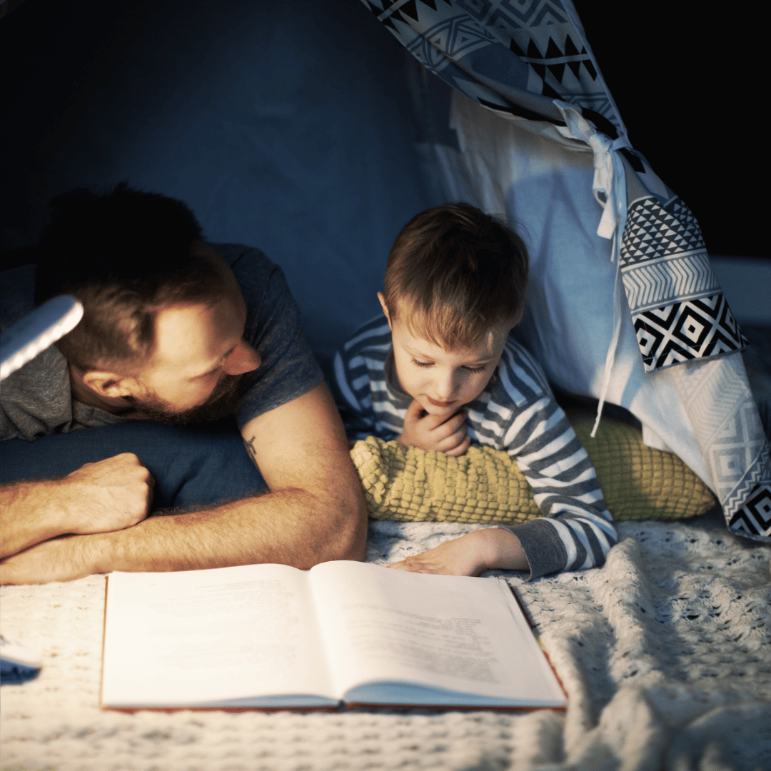 A man and a young boy lie under a cozy blanket inside an Elementor-inspired tent, reading a book together by the gentle glow of a small lamp. Both appear engaged and comfortable in the warm, softly lit setting.