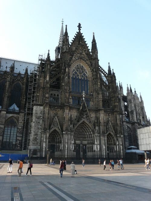 The intricate facade of St. Vitus Cathedral, showcasing detailed gothic architecture with tall spires and ornate stonework, surrounded by people walking in a plaza under a clear blue sky.