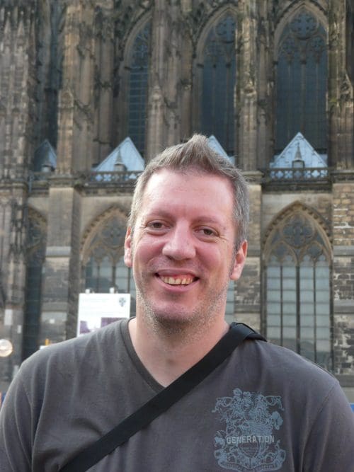 A smiling man in a gray shirt stands in front of a large, ornate stone building with tall gothic windows and intricate architectural details.