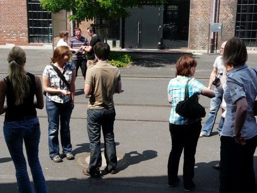 A group of people gathered on a sunny street, engaged in conversation. They stand on a paved surface, surrounded by brick buildings and greenery, with some individuals holding papers or smartphones.