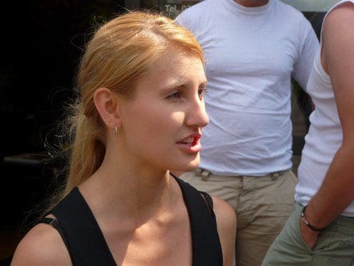 A woman with long blonde hair, wearing a sleeveless black top, is engaged in conversation. She has hoop earrings and a thoughtful expression. Two people in casual clothing are slightly blurred in the background.