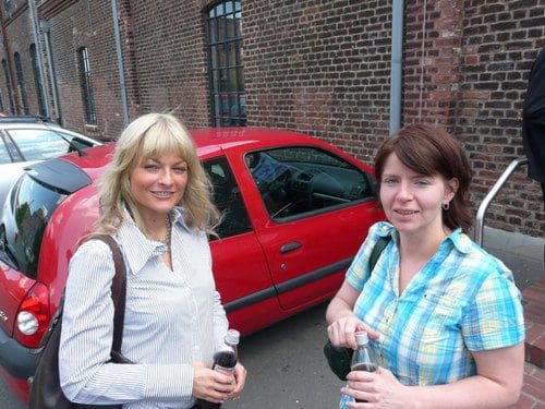 Two women standing outside near a red car parked beside a brick building. One woman has long blonde hair and holds a drink, while the other has shoulder-length brown hair, also holding a beverage. They are both casually dressed.