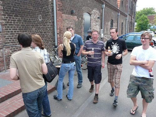 A group of young adults gathered on a street, some walking and others conversing. They are dressed casually, with drinks in hand, and surrounded by brick buildings and bicycles parked nearby.