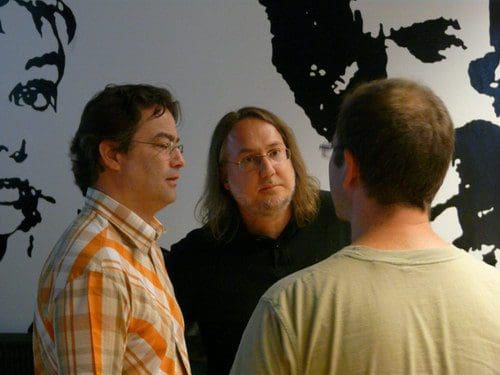 Three men engaged in conversation, standing close together in front of a wall featuring artistic black-and-white portraits. One man wears a striped orange and white shirt, another has long hair and glasses, and the third has a short haircut.