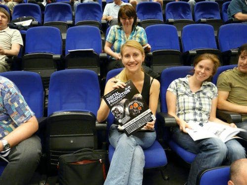 A group of people seated in an auditorium, with a woman in the foreground holding a flyer. She has long blonde hair and is wearing a black top. Other attendees are visible in the background, engaged in conversation.