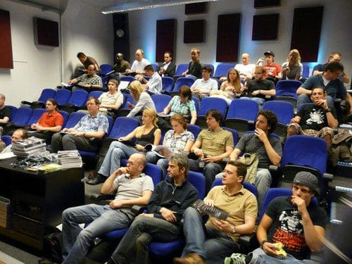 An audience of diverse individuals seated in a modern lecture hall with blue chairs, attentively watching a presentation. Some attendees are taking notes while others are engaged in conversation.