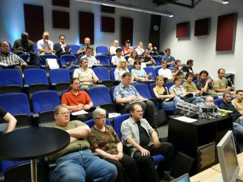 An audience of diverse individuals sitting in a lecture hall with blue seating. Some attendees are focused on their devices, while others are attentively watching a presentation, with a mixture of expressions across their faces.