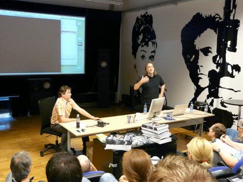 A presenter gesturing while speaking to an audience in a modern classroom. A large screen displays content behind them, and a table is set with laptops and photography equipment. A striking mural of a figure is painted on the wall.