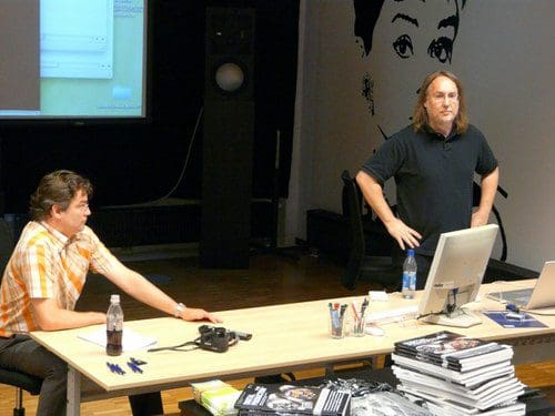 Two men participating in a discussion at a table in a modern setting. One man, wearing a striped shirt, sits with a drink and camera, while another man stands beside a computer, gesturing towards a screen in the background.