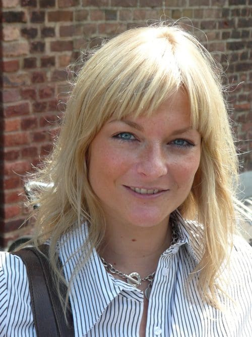 A woman with long blonde hair and blue eyes smiling while wearing a striped shirt. The background features a brick wall, suggesting an outdoor setting.