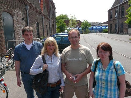 A group of four people standing on a sunny day outside a brick building. They are casually dressed, with two men and two women, and there are bicycles parked nearby. The background includes trees and a street.