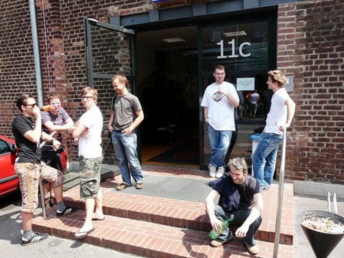 A group of six men gathered outside a building marked '11c'. They are casually dressed, some standing on steps and others sitting. The scene is set against a brick wall on a sunny day, with a few parked cars in the background.