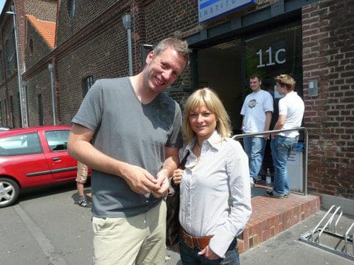 Two people smiling outside a building with a sign labeled '11c.' The man is wearing a gray t-shirt and khaki shorts, while the woman wears a white button-up shirt and blue jeans. A red car is parked nearby, and others are seen in the background.