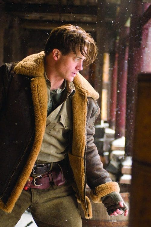 A young man wearing a brown shearling-lined jacket and a green shirt stands at an old, wooden structure. Flurries of snow fall around him, creating a chilly atmosphere as he gazes thoughtfully into the distance.