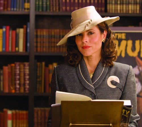 A woman in a vintage gray suit and a large cream hat stands in front of a lectern, holding papers. Behind her is a bookshelf filled with colorful books, creating an academic atmosphere.