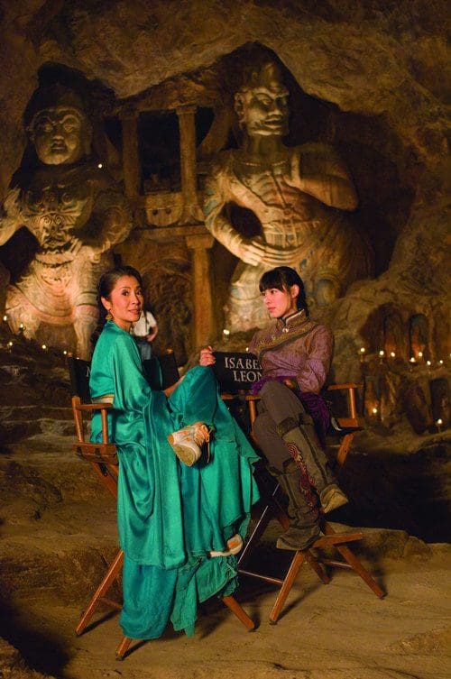Two women seated in a dimly lit cave with intricate stone sculptures in the background. One woman wears a flowing green outfit while the other is dressed in a purple and gray attire, both engaged in conversation.